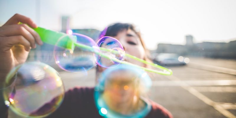 Mile Two: Creativity. Image is a close-up of young lady wearing a warm-looking purple sweater blowing bubbles into the camera. She is standing on what appears to be the top of a parking garage, and even though it looks chilly outside, a lens flare in the background reveals that it is at least a sunny day. 